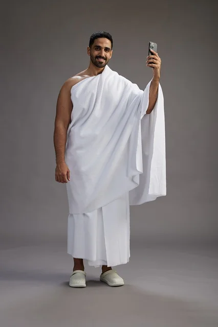 بورتريه لرجل سعودي بملابس الإحرام يبتسم ويلتقط صورة سيلفي, استخدام الهاتف المحمول, الحج والعمره في مكه, الجسم بالكامل, خلفيه رماديه