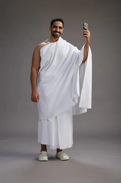 A portrait of a Saudi man in Ihram clothing smiling and taking a selfie, using a mobile phone, during Hajj and Umrah in Mecca, full body, gray background.