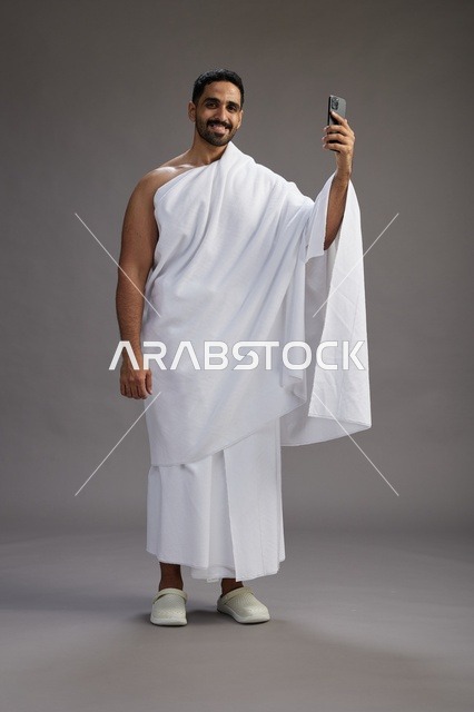 بورتريه لرجل سعودي بملابس الإحرام يبتسم ويلتقط صورة سيلفي, استخدام الهاتف المحمول, الحج والعمره في مكه, الجسم بالكامل, خلفيه رماديه