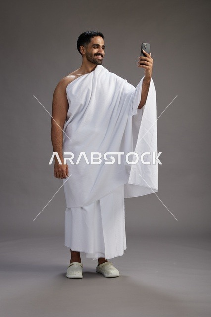 بورتريه لرجل سعودي بملابس الإحرام يبتسم ويلتقط صورة سيلفي, استخدام الهاتف المحمول, الحج والعمره في مكه, الجسم بالكامل, خلفيه رماديه