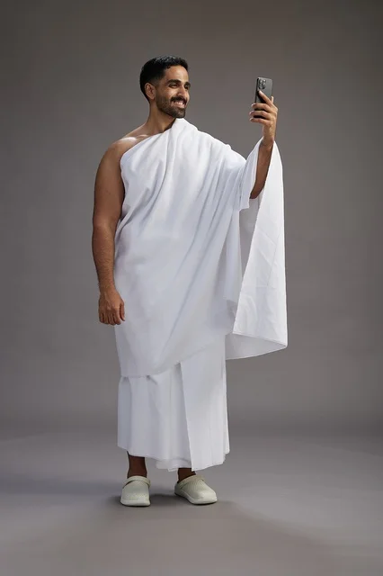 بورتريه لرجل سعودي بملابس الإحرام يبتسم ويلتقط صورة سيلفي, استخدام الهاتف المحمول, الحج والعمره في مكه, الجسم بالكامل, خلفيه رماديه
