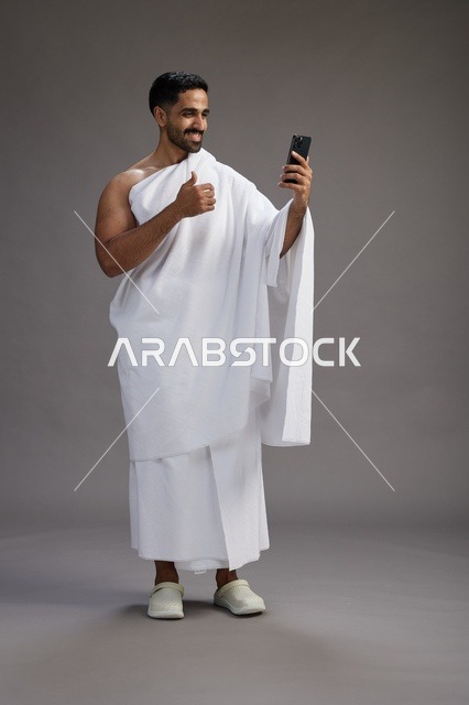 بورتريه لرجل سعودي بملابس الإحرام يبتسم ويلتقط صورة سيلفي, استخدام الهاتف المحمول, الحج والعمره في مكه, الجسم بالكامل, خلفيه رماديه
