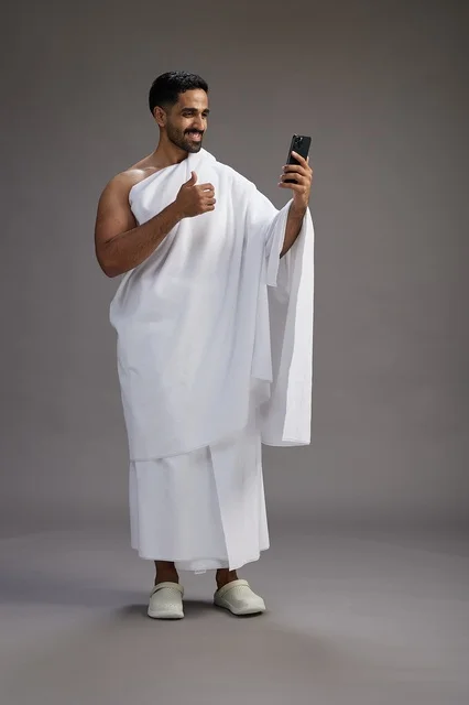 بورتريه لرجل سعودي بملابس الإحرام يبتسم ويلتقط صورة سيلفي, استخدام الهاتف المحمول, الحج والعمره في مكه, الجسم بالكامل, خلفيه رماديه