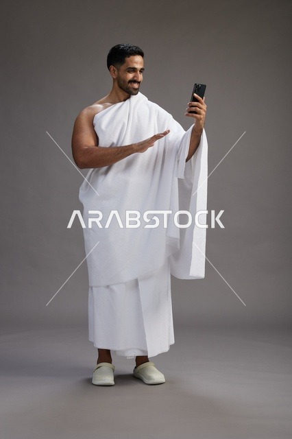 بورتريه لرجل سعودي بملابس الإحرام يبتسم ويلتقط صورة سيلفي, استخدام الهاتف المحمول, الحج والعمره في مكه, الجسم بالكامل, خلفيه رماديه