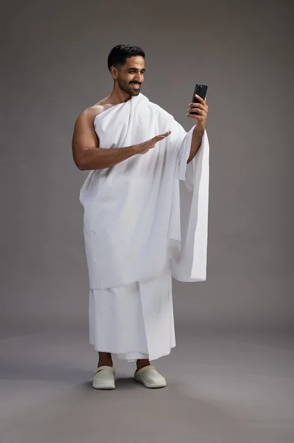 بورتريه لرجل سعودي بملابس الإحرام يبتسم ويلتقط صورة سيلفي, استخدام الهاتف المحمول, الحج والعمره في مكه, الجسم بالكامل, خلفيه رماديه