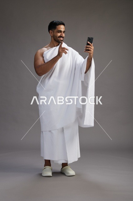 بورتريه لرجل سعودي بملابس الإحرام يبتسم ويلتقط صورة سيلفي, استخدام الهاتف المحمول, الحج والعمره في مكه, الجسم بالكامل, خلفيه رماديه