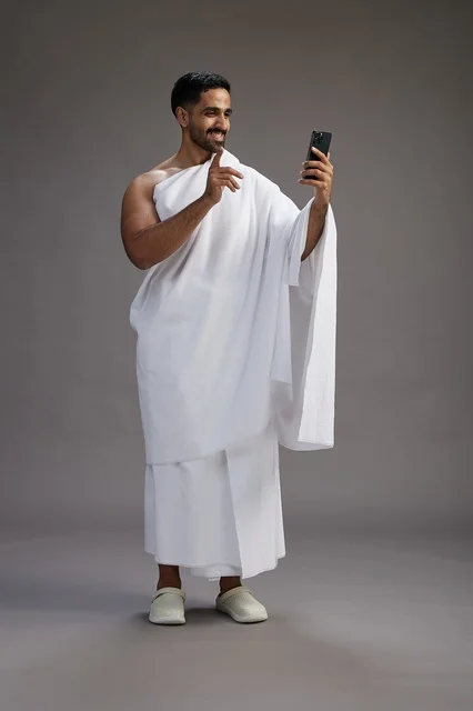 بورتريه لرجل سعودي بملابس الإحرام يبتسم ويلتقط صورة سيلفي, استخدام الهاتف المحمول, الحج والعمره في مكه, الجسم بالكامل, خلفيه رماديه