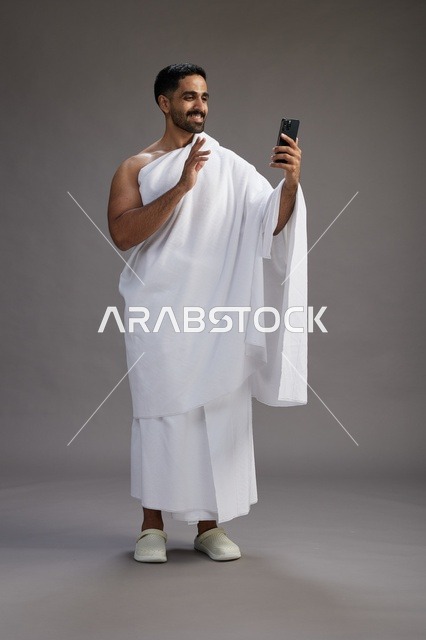بورتريه لرجل سعودي بملابس الإحرام يبتسم ويلتقط صورة سيلفي, استخدام الهاتف المحمول, الحج والعمره في مكه, الجسم بالكامل, خلفيه رماديه