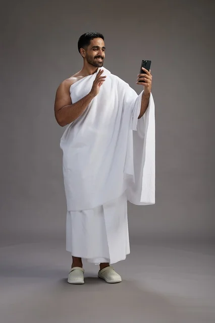بورتريه لرجل سعودي بملابس الإحرام يبتسم ويلتقط صورة سيلفي, استخدام الهاتف المحمول, الحج والعمره في مكه, الجسم بالكامل, خلفيه رماديه
