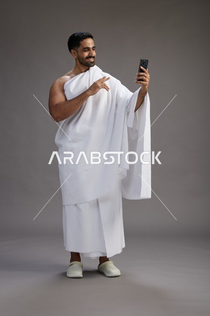 بورتريه لرجل سعودي بملابس الإحرام يبتسم ويلتقط صورة سيلفي, استخدام الهاتف المحمول, الحج والعمره في مكه, الجسم بالكامل, خلفيه رماديه