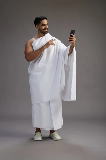 A portrait of a Saudi man in Ihram clothing smiling and taking a selfie, using a mobile phone, during Hajj and Umrah in Mecca, full body, gray background.
