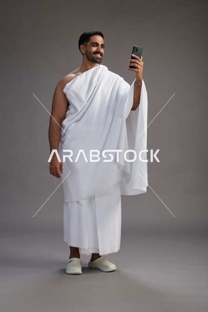 بورتريه لرجل سعودي بملابس الإحرام يبتسم ويلتقط صورة سيلفي, استخدام الهاتف المحمول, الحج والعمره في مكه, الجسم بالكامل, خلفيه رماديه