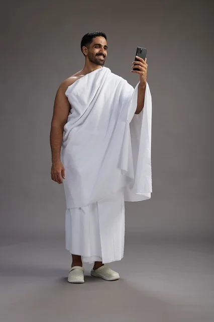 A portrait of a Saudi man in Ihram clothing smiling and taking a selfie, using a mobile phone, during Hajj and Umrah in Mecca, full body, gray background.