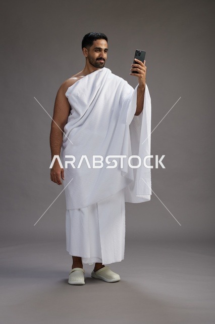 بورتريه لرجل سعودي بملابس الإحرام يبتسم ويلتقط صورة سيلفي, استخدام الهاتف المحمول, الحج والعمره في مكه, الجسم بالكامل, خلفيه رماديه