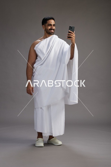 بورتريه لرجل سعودي بملابس الإحرام يبتسم ويلتقط صورة سيلفي, استخدام الهاتف المحمول, الحج والعمره في مكه, الجسم بالكامل, خلفيه رماديه