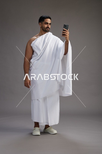 بورتريه لرجل سعودي بملابس الإحرام يبتسم ويلتقط صورة سيلفي, استخدام الهاتف المحمول, الحج والعمره في مكه, الجسم بالكامل, خلفيه رماديه
