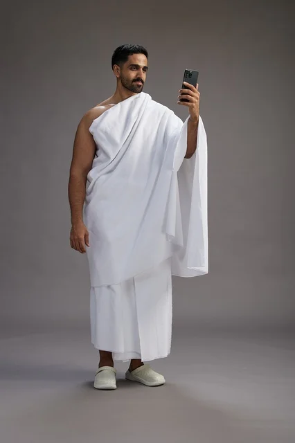 A portrait of a Saudi man in Ihram clothing smiling and taking a selfie, using a mobile phone, during Hajj and Umrah in Mecca, full body, gray background.