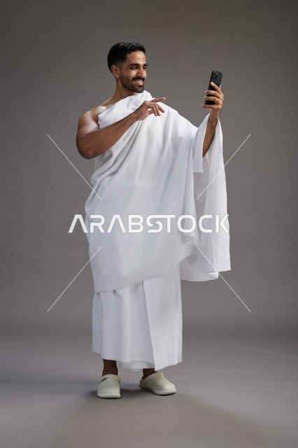 بورتريه لرجل سعودي بملابس الإحرام يبتسم ويلتقط صورة سيلفي, استخدام الهاتف المحمول, الحج والعمره في مكه, الجسم بالكامل, خلفيه رماديه