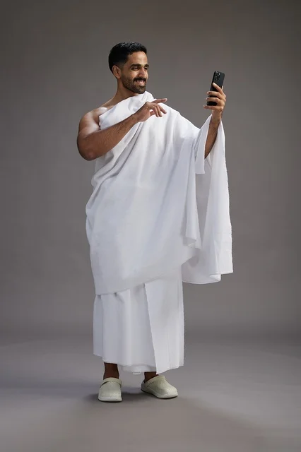 بورتريه لرجل سعودي بملابس الإحرام يبتسم ويلتقط صورة سيلفي, استخدام الهاتف المحمول, الحج والعمره في مكه, الجسم بالكامل, خلفيه رماديه