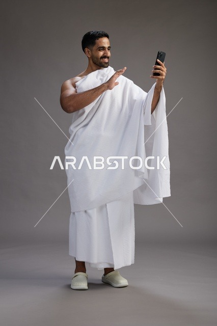 بورتريه لرجل سعودي بملابس الإحرام يبتسم ويلتقط صورة سيلفي, استخدام الهاتف المحمول, الحج والعمره في مكه, الجسم بالكامل, خلفيه رماديه