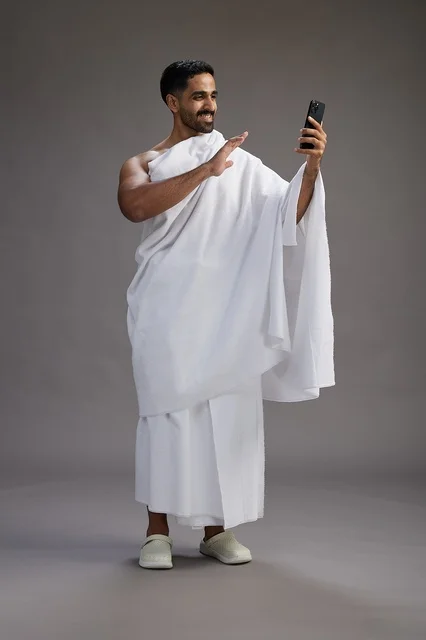 A portrait of a Saudi man in Ihram clothing smiling and taking a selfie, using a mobile phone, during Hajj and Umrah in Mecca, full body, gray background.
