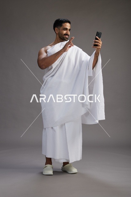 بورتريه لرجل سعودي بملابس الإحرام يبتسم ويلتقط صورة سيلفي, استخدام الهاتف المحمول, الحج والعمره في مكه, الجسم بالكامل, خلفيه رماديه