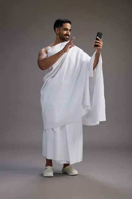 A portrait of a Saudi man in Ihram clothing smiling and taking a selfie, using a mobile phone, during Hajj and Umrah in Mecca, full body, gray background.
