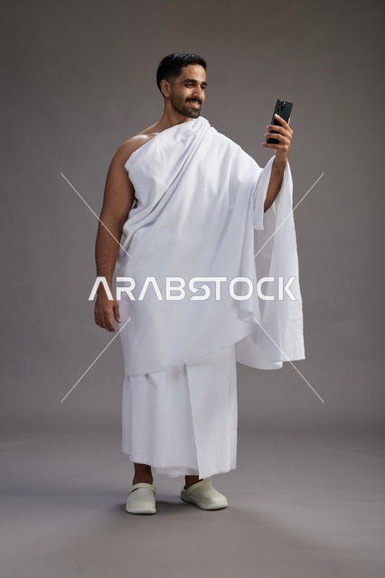 بورتريه لرجل سعودي بملابس الإحرام يبتسم ويلتقط صورة سيلفي, استخدام الهاتف المحمول, الحج والعمره في مكه, الجسم بالكامل, خلفيه رماديه