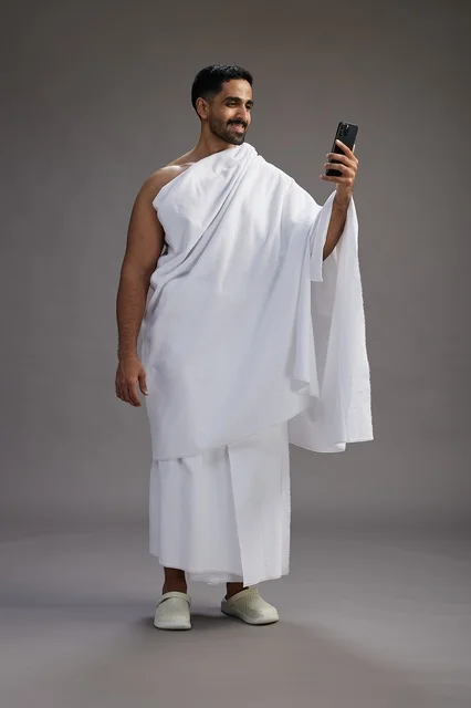 A portrait of a Saudi man in Ihram clothing smiling and taking a selfie, using a mobile phone, during Hajj and Umrah in Mecca, full body, gray background.