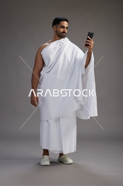 بورتريه لرجل سعودي بملابس الإحرام يبتسم ويلتقط صورة سيلفي, استخدام الهاتف المحمول, الحج والعمره في مكه, الجسم بالكامل, خلفيه رماديه