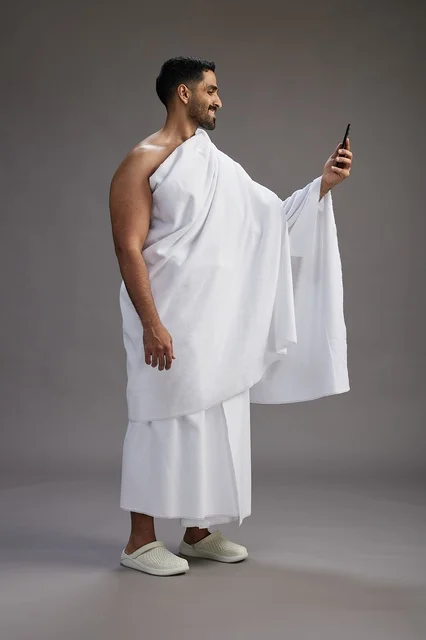 بورتريه لرجل سعودي بملابس الإحرام يبتسم ويلتقط صورة سيلفي, استخدام الهاتف المحمول, الحج والعمره في مكه, الجسم بالكامل, خلفيه رماديه