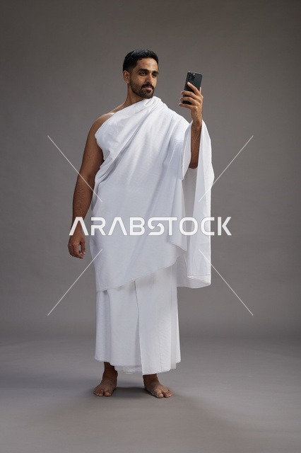 بورتريه لرجل سعودي بملابس الإحرام يبتسم ويلتقط صورة سيلفي, استخدام الهاتف المحمول, الحج والعمره في مكه, الجسم بالكامل, خلفيه رماديه