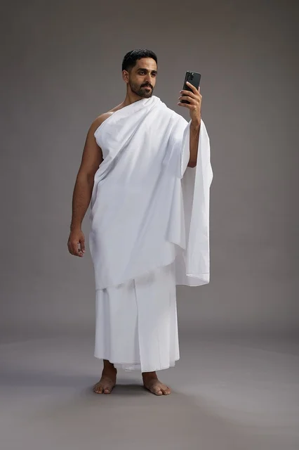 A portrait of a Saudi man in Ihram clothing smiling and taking a selfie, using a mobile phone, during Hajj and Umrah in Mecca, full body, gray background.