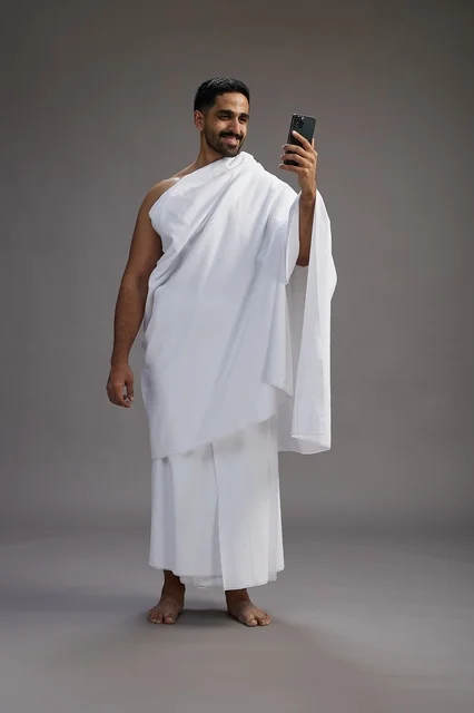 A portrait of a Saudi man in Ihram clothing smiling and taking a selfie, using a mobile phone, during Hajj and Umrah in Mecca, full body, gray background.