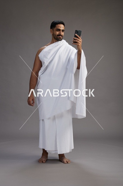 بورتريه لرجل سعودي بملابس الإحرام يبتسم ويلتقط صورة سيلفي, استخدام الهاتف المحمول, الحج والعمره في مكه, الجسم بالكامل, خلفيه رماديه