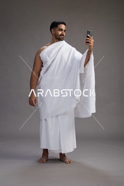 بورتريه لرجل سعودي بملابس الإحرام يبتسم ويلتقط صورة سيلفي, استخدام الهاتف المحمول, الحج والعمره في مكه, الجسم بالكامل, خلفيه رماديه
