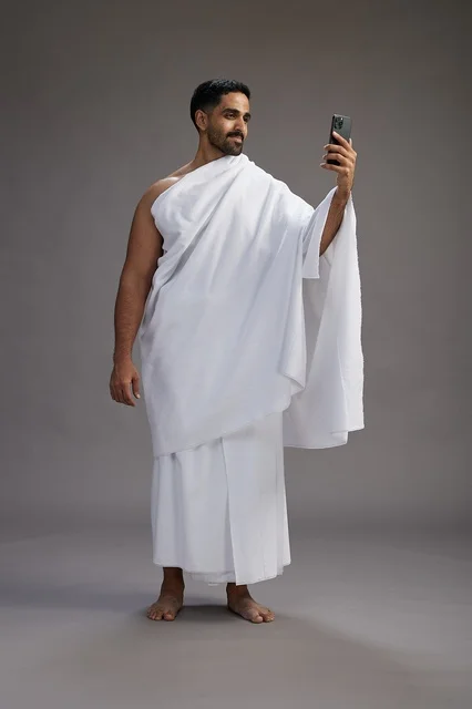بورتريه لرجل سعودي بملابس الإحرام يبتسم ويلتقط صورة سيلفي, استخدام الهاتف المحمول, الحج والعمره في مكه, الجسم بالكامل, خلفيه رماديه