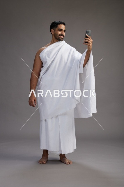 بورتريه لرجل سعودي بملابس الإحرام يبتسم ويلتقط صورة سيلفي, استخدام الهاتف المحمول, الحج والعمره في مكه, الجسم بالكامل, خلفيه رماديه