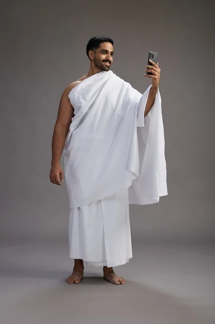 A portrait of a Saudi man in Ihram clothing smiling and taking a selfie, using a mobile phone, during Hajj and Umrah in Mecca, full body, gray background.