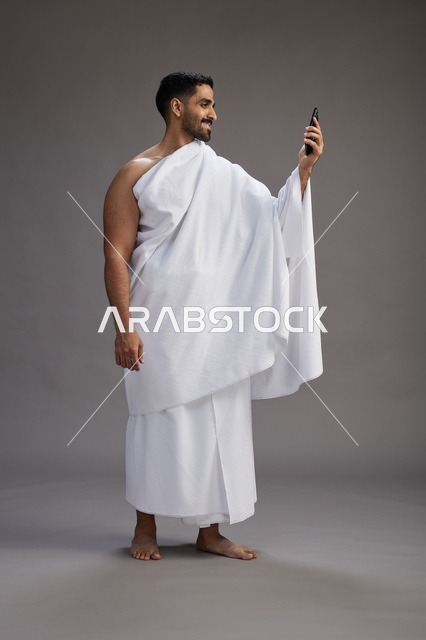 بورتريه لرجل سعودي بملابس الإحرام يبتسم ويلتقط صورة سيلفي, استخدام الهاتف المحمول, الحج والعمره في مكه, الجسم بالكامل, خلفيه رماديه