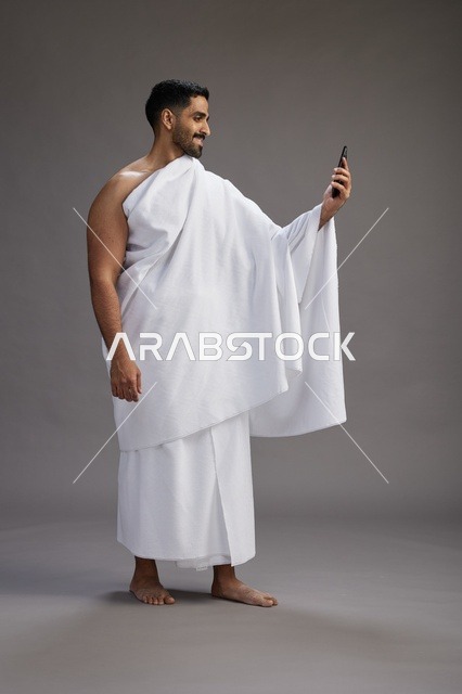 بورتريه لرجل سعودي بملابس الإحرام يبتسم ويلتقط صورة سيلفي, استخدام الهاتف المحمول, الحج والعمره في مكه, الجسم بالكامل, خلفيه رماديه