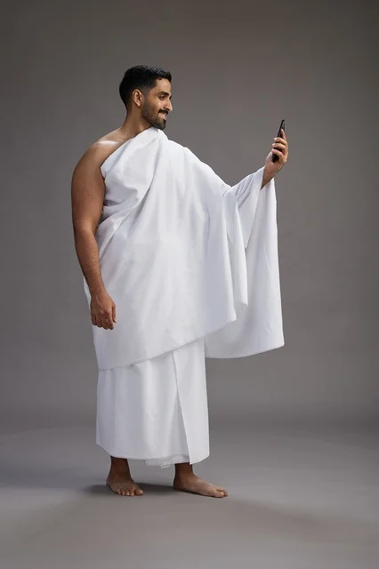 A portrait of a Saudi man in Ihram clothing smiling and taking a selfie, using a mobile phone, during Hajj and Umrah in Mecca, full body, gray background.