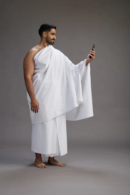 A portrait of a Saudi man in Ihram clothing smiling and taking a selfie, using a mobile phone, during Hajj and Umrah in Mecca, full body, gray background.