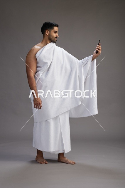 A portrait of a Saudi man in Ihram clothing smiling and taking a selfie, using a mobile phone, during Hajj and Umrah in Mecca, full body, gray background.