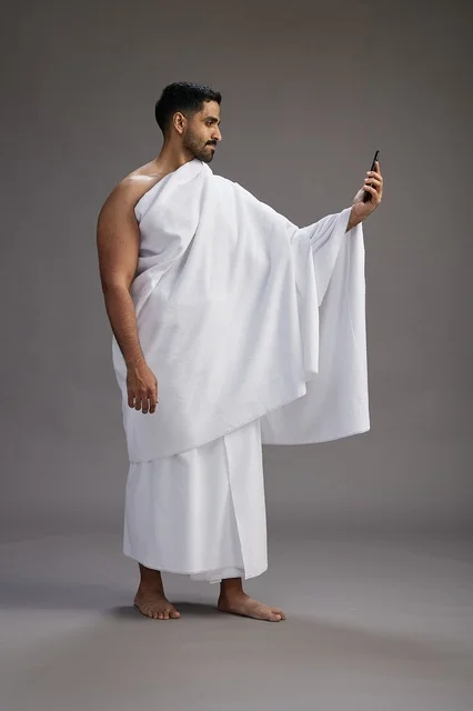 بورتريه لرجل سعودي بملابس الإحرام يبتسم ويلتقط صورة سيلفي, استخدام الهاتف المحمول, الحج والعمره في مكه, الجسم بالكامل, خلفيه رماديه