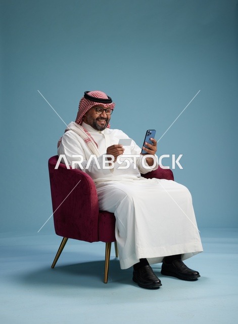 بورتريه لرجل سعودي يستخدم الهاتف المحمول يجلس على كرسي وهو يحمل بطاقة بيضاء فارغه,  استخدام البطاقه البنكيه, خلفيه زرقاء