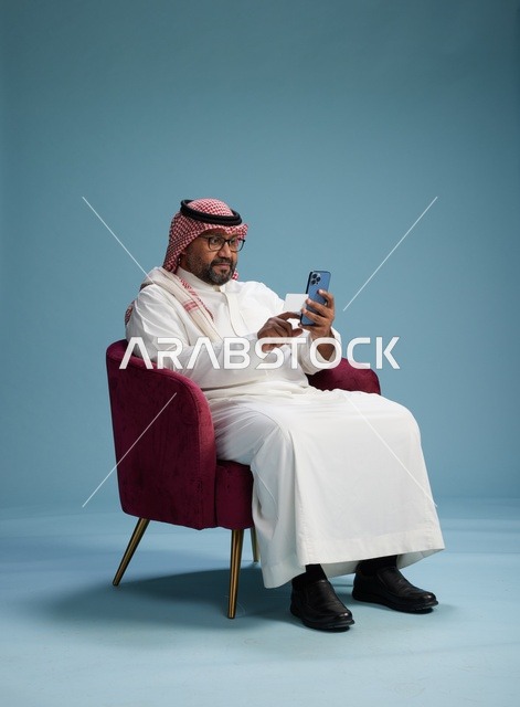 بورتريه لرجل سعودي يستخدم الهاتف المحمول يجلس على كرسي وهو يحمل بطاقة بيضاء فارغه,  استخدام البطاقه البنكيه, خلفيه زرقاء