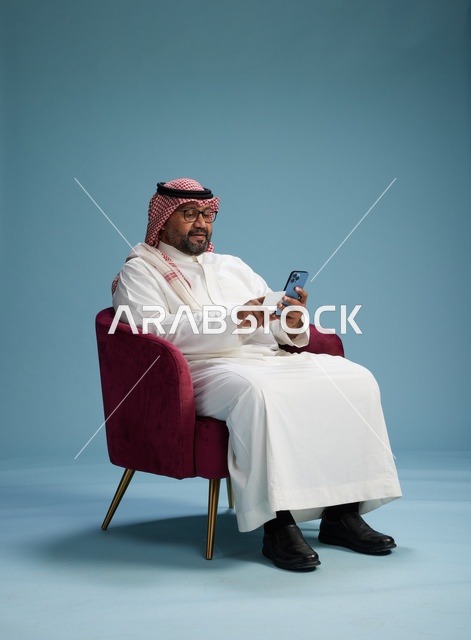 بورتريه لرجل سعودي يستخدم الهاتف المحمول يجلس على كرسي وهو يحمل بطاقة بيضاء فارغه,  استخدام البطاقه البنكيه, خلفيه زرقاء