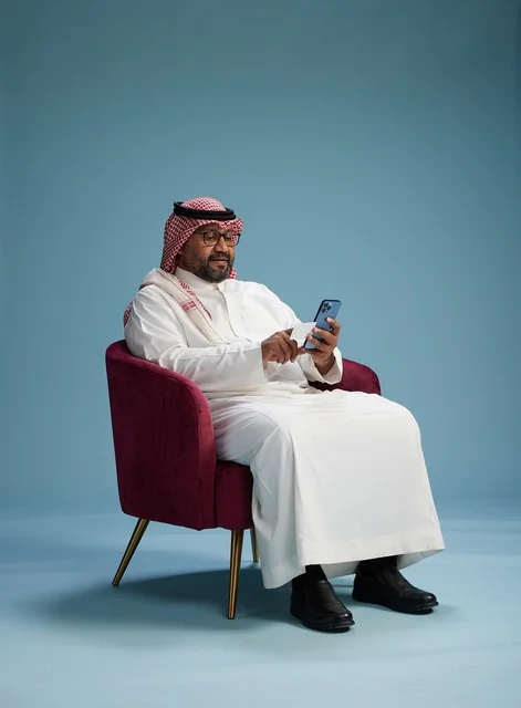 A portrait of a Saudi man using a mobile phone sitting on a chair while holding a blank white card, using a bank card, with a blue background.