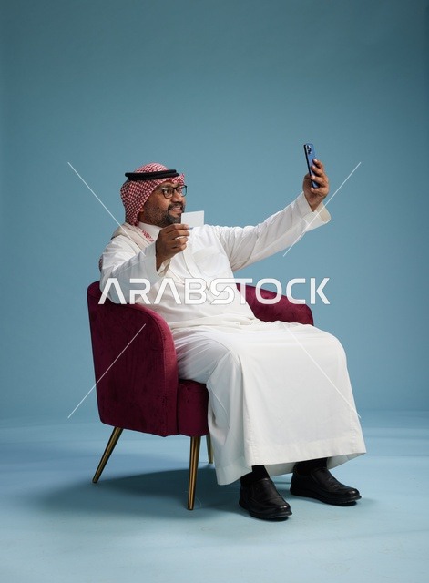 بورتريه لرجل سعودي يستخدم الهاتف المحمول يجلس على كرسي وهو يحمل بطاقة بيضاء فارغه,  استخدام البطاقه البنكيه, خلفيه زرقاء